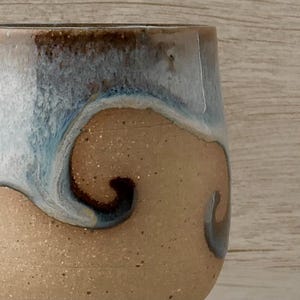 May include: Close-up of a handmade ceramic tumbler. The cup features a speckled tan base with a swirling design of blue, white, and brown glaze. The rim is a darker brown. The design gives the cup a unique, artistic look.
