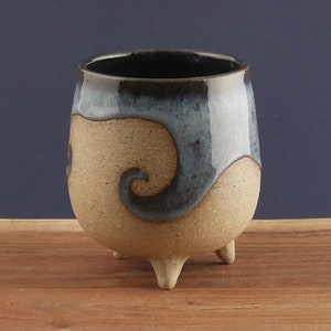 May include: A handmade ceramic cup with a unique design. The cup features a speckled tan base, a dark blue and black glazed rim, and a decorative swirl. It stands on three small feet, showcasing a blend of rustic and modern aesthetics.