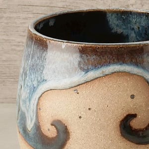 May include: A handmade ceramic mug with a unique design. The mug features a tan base with dark brown and blue accents. The rim is dark brown, and the interior is a deep blue. The mug has a smooth, glossy finish.
