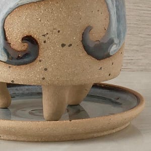 May include: A handmade ceramic planter with a speckled tan exterior and a blue and white wave design. The planter sits on three short legs and includes a matching saucer. The design is a unique piece of pottery.