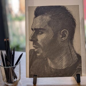May include: A graphite pencil drawing on paper depicting a profile view of a person with short hair and facial stubble. The artwork is displayed on a small black stand next to a clear glass container filled with several black drawing pencils.