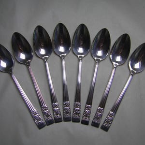 May include: A collection of seven silver spoons, fanned out. Each spoon features a polished, oval bowl and a long, slender handle with a floral design near the base. The spoons' surfaces reflect the surroundings, creating a mirrored effect.