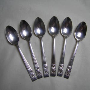 May include: Six silver teaspoons with ornate designs on the handles. The spoons have a polished finish and are arranged in a row.