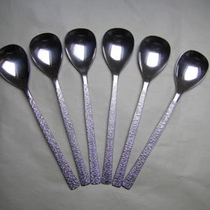 May include: Six stainless steel soup spoons. The spoons are arranged in a fan shape. They have textured handles and shiny, reflective bowls. The spoons are silver in colour.