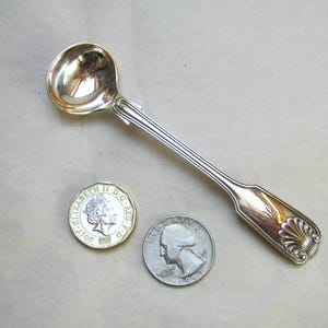 May include: A silver-toned metal spoon with a round bowl and a handle featuring a ribbed design and a shell motif at the end. The spoon is positioned next to a 2017 British one pound coin and a 1967 American quarter dollar for scale reference.