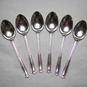 May include: Six silver spoons arranged in a fan shape. The spoons have oval bowls and long, slender handles with a decorative beaded pattern near the bowl. The spoons are reflective, showing the surrounding environment.