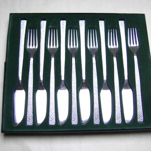 May include: A set of eight silver-toned metal fish forks arranged in a rectangular dark green display box. Each fork features a polished tined head and a textured, hammered-style handle, presented in individual moulded slots within the container.