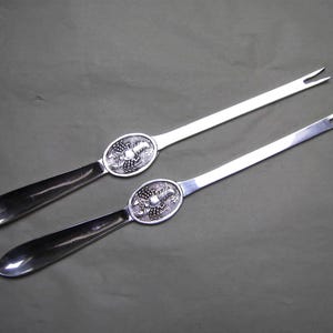 May include: Two silver serving forks with a decorative oval center featuring a detailed design. The handles are dark and polished, and the forks have two tines. The background is a muted green.