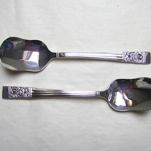 May include: Two silver-toned metal spoons with wide, rounded bowls and handles featuring a decorative embossed floral pattern at the end. The handles have a series of vertical ridges leading from the bowl to the detailed floral design.