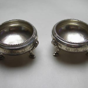 May include: Two silver salt cellars with three legs each. The cellars have a beaded rim and a decorative pattern on the bottom.