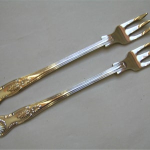 May include: Two silver-plated forks with ornate handles. The forks have a gold finish on the handles and are designed for serving desserts or appetizers.