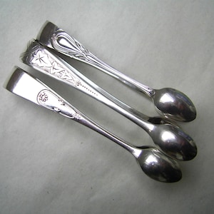 May include: Three silver sugar spoons with decorative handles. One spoon has a floral design, another a starburst pattern, and the third a heart-shaped design. The spoons are displayed on a light-coloured surface.