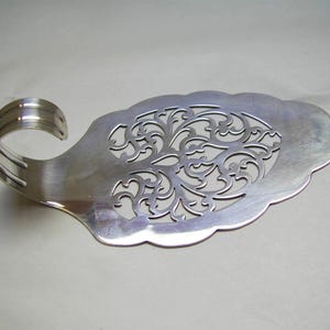 May include: A silver cake server with a decorative handle and a scalloped edge. The server has an openwork floral design in the centre. The handle has a curved design. The server is made of metal.