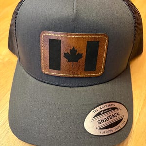 Peut inclure: Une casquette de camionneur en maille grise et noire avec un patch en cuir brun représentant le drapeau canadien. La casquette a un dos en maille noire et un devant gris avec une visière cousue. Le patch a une bordure en cuir brun et un drapeau canadien en cuir noir avec une feuille d'érable brune. La casquette a une étiquette argentée au dos qui indique "The Authentic Yupoong Snapback Yupoong Since 1974."