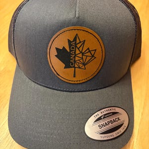 May include: A grey and black mesh trucker hat with a brown leather patch featuring a black maple leaf and the word "CANADA". The hat has a silver label that reads "THE AUTHENTIC YUPOONG SNAPBACK YUPOONG SINCE 1974".