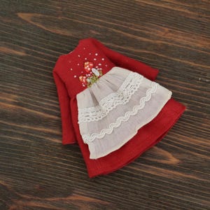 May include: A red linen doll dress with embroidered mushrooms and a white lace trim.