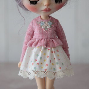 May include: A pink doll wearing a pink and white floral embroidered top and a white skirt with a floral print and lace trim.