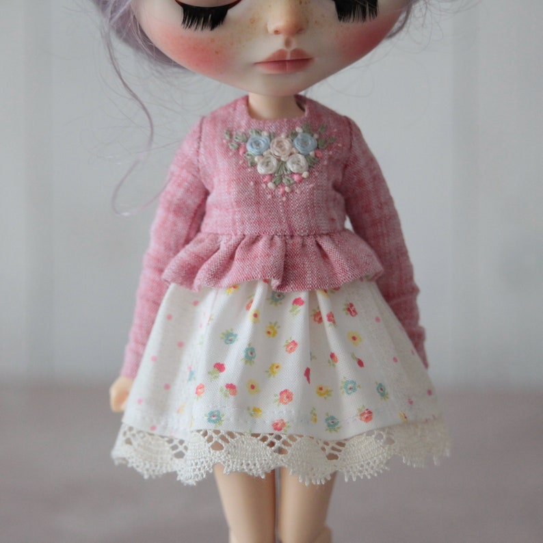 May include: A pink doll wearing a pink and white floral embroidered top and a white skirt with a floral print and lace trim.