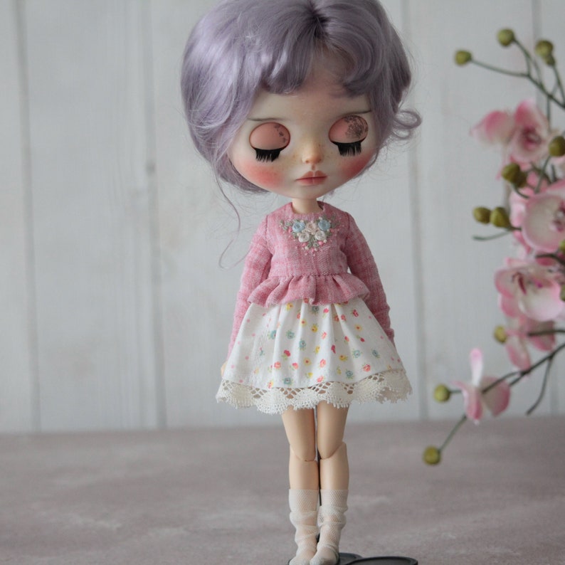 May include: A doll with lavender hair wearing a pink ruffled sweater with floral embroidery and a white skirt with a floral print and lace trim.