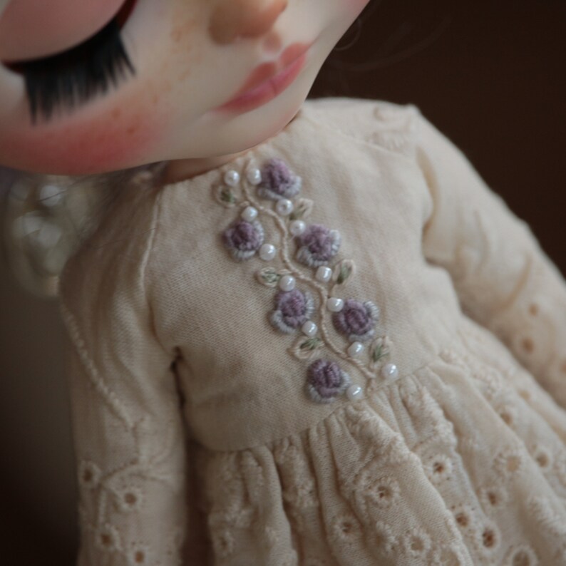 May include: A white doll's dress with a delicate floral embroidery design featuring purple flowers and white pearls.
