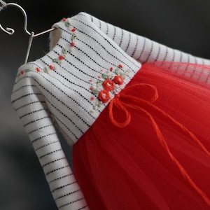 May include: A miniature dress with a white and black striped bodice and a bright red tulle skirt. The bodice is adorned with embroidered floral details. The dress is hanging on a silver hanger.