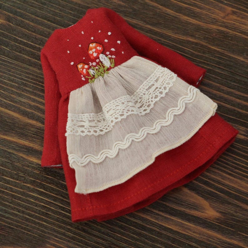 May include: A red linen dress with embroidered mushrooms and white lace trim. The dress has long sleeves and a gathered skirt.