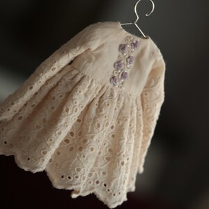 May include: A delicate white eyelet dress with long sleeves and a floral embroidery design on the bodice. The dress has a scalloped hemline.