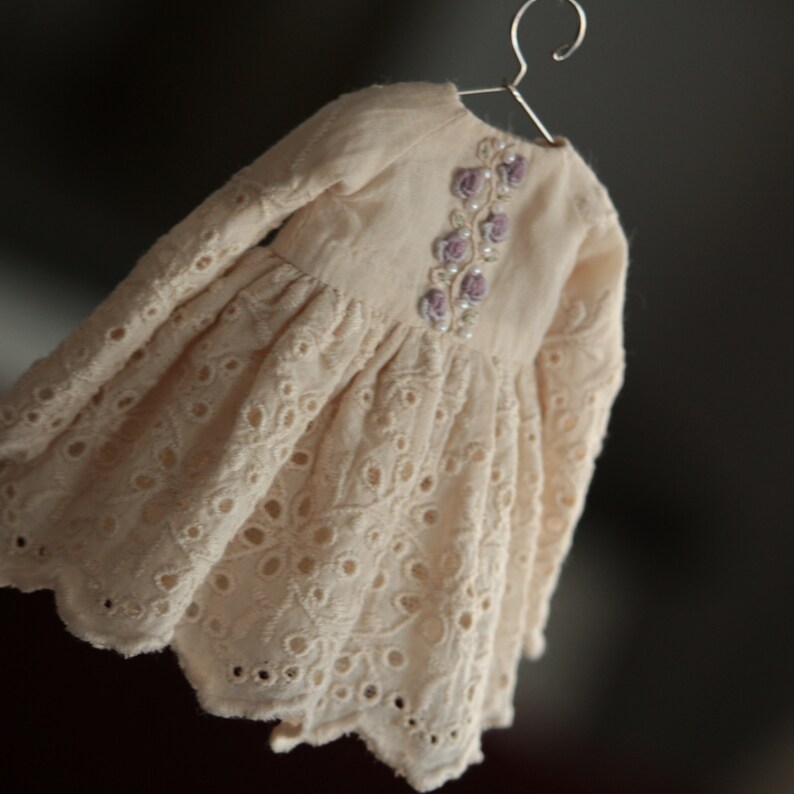 May include: A delicate white eyelet dress with long sleeves and a floral embroidery design on the bodice. The dress has a scalloped hemline.