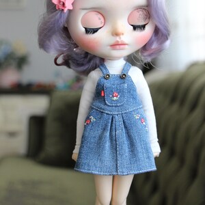 May include: A doll with closed eyes, wearing a white long-sleeved shirt and a denim overall dress. The dress features embroidered floral details and a small pocket. The doll has purple hair and a pink flower hair accessory.