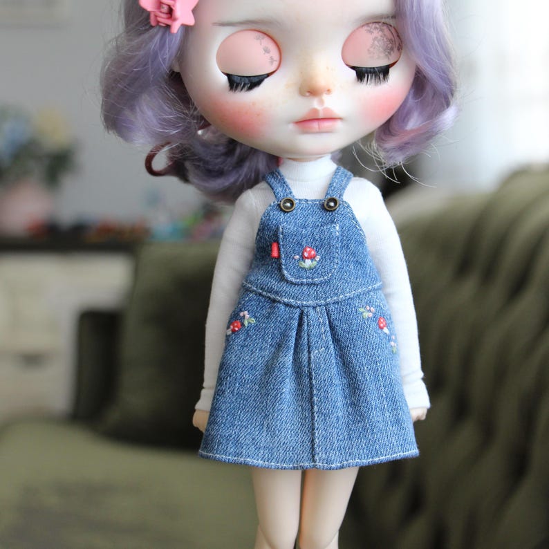 May include: A doll with closed eyes, wearing a white long-sleeved shirt and a denim overall dress. The dress features embroidered floral details and a small pocket. The doll has purple hair and a pink flower hair accessory.