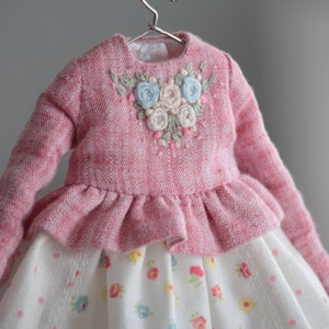 May include: A pink knitted dress with a ruffled skirt and a floral embroidered bodice. The dress has a white and floral print skirt.