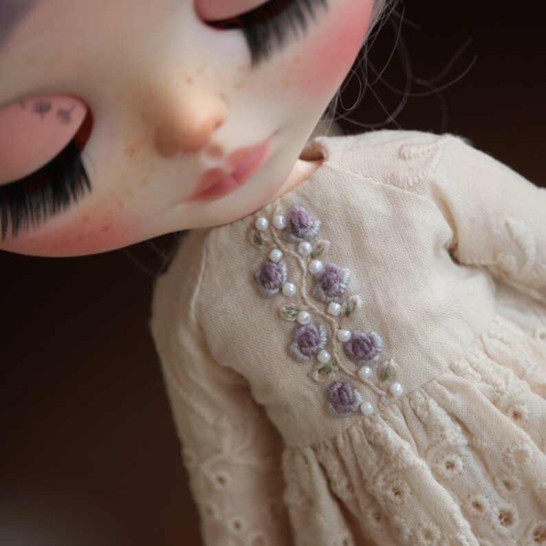 May include: A close-up of a doll wearing a cream-coloured dress with delicate embroidery of purple flowers and white pearls.
