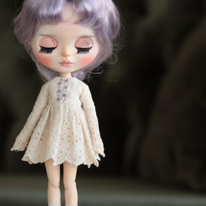 May include: A doll with purple hair wearing a white eyelet dress with a beaded bodice. The doll has painted eyelashes and blush.