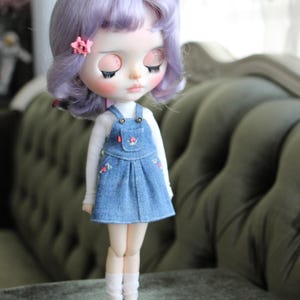 May include: A doll with lavender hair and a pink flower hair clip. The doll wears a white long-sleeved shirt under a denim overall dress with floral embroidery. The doll has closed eyes and is wearing white socks.