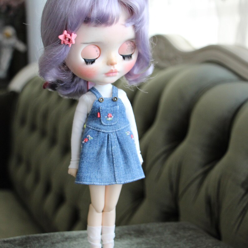 May include: A doll with lavender hair and a pink flower hair clip. The doll wears a white long-sleeved shirt under a denim overall dress with floral embroidery. The doll has closed eyes and is wearing white socks.
