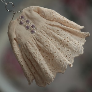 May include: A delicate, off-white dress with long sleeves and a scalloped hem. The dress features intricate eyelet lace detailing and a floral embroidery design with purple flowers and pearl accents.