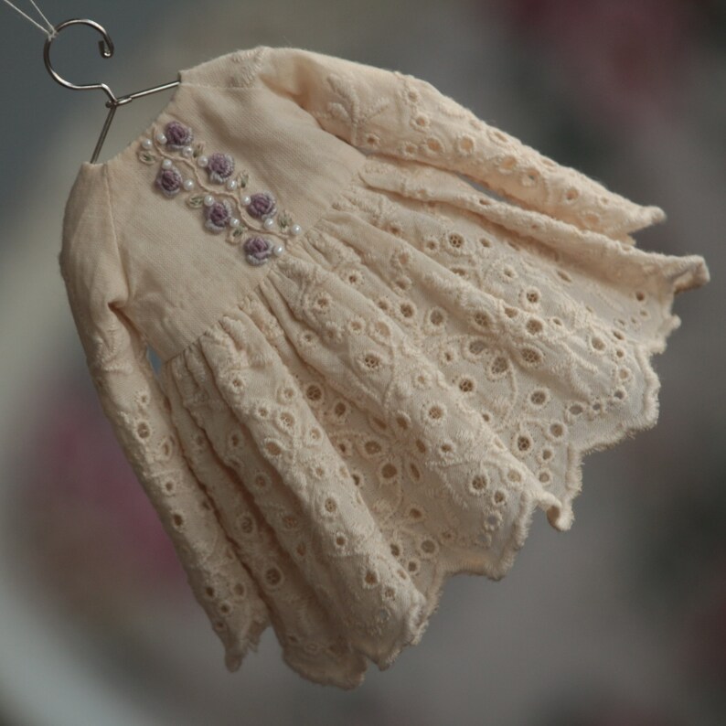 May include: A delicate, off-white dress with long sleeves and a scalloped hem. The dress features intricate eyelet lace detailing and a floral embroidery design with purple flowers and pearl accents.
