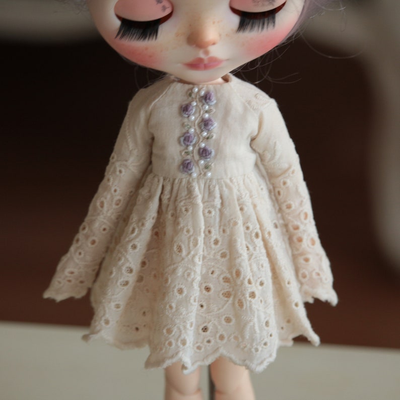 May include: A white eyelet dress with a floral embroidery detail on the bodice. The dress has long sleeves and a scalloped hem.