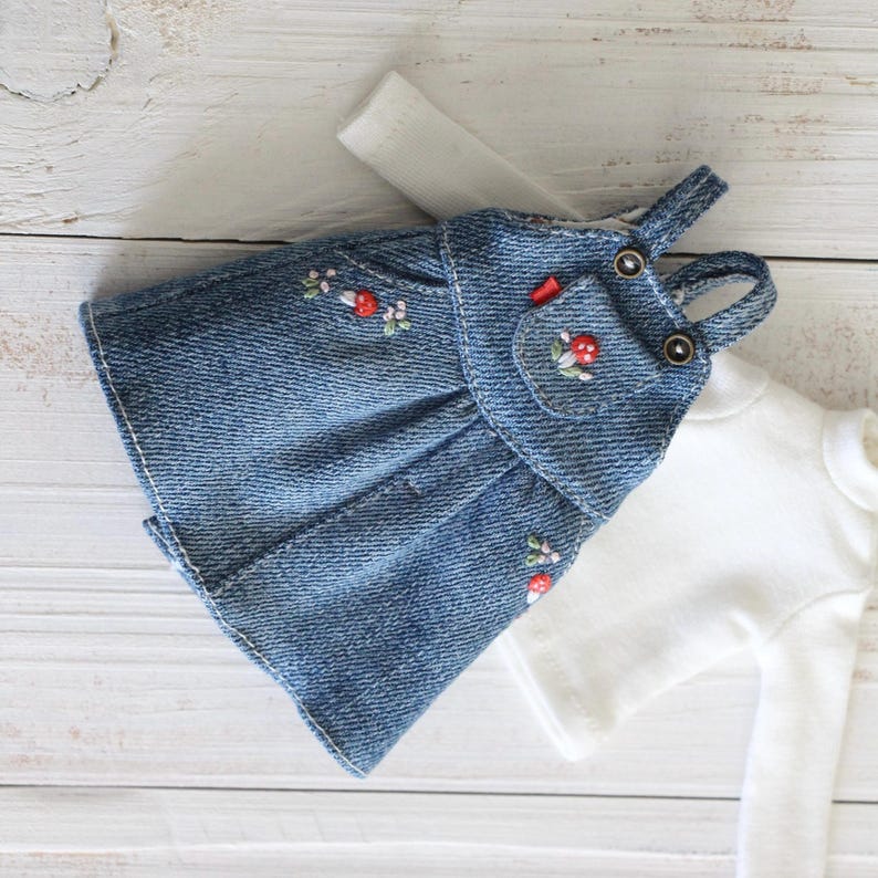 May include: A miniature denim dress with embroidered red flowers and a small pocket. The dress has adjustable straps and is paired with a white long-sleeved shirt. The outfit is designed for a doll.