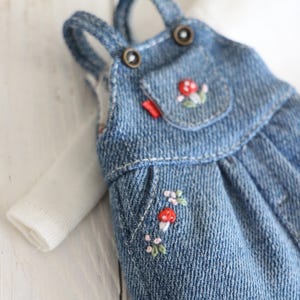 May include: A miniature denim overall dress with embroidered mushroom and floral details. The blue jean dress has a small pocket and red tag. A white long-sleeved shirt is visible underneath. The overall straps are secured with bronze-colored buttons.