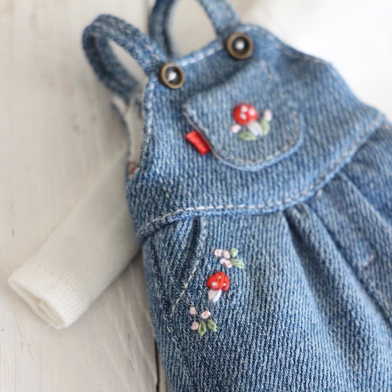 May include: A miniature denim overall dress with embroidered mushroom and floral details. The blue jean dress has a small pocket and red tag. A white long-sleeved shirt is visible underneath. The overall straps are secured with bronze-colored buttons.