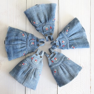 May include: Five denim dresses with embroidered floral details are arranged in a circular pattern. Each dress features a bib pocket and adjustable straps. The dresses are a light blue color and are set against a white wooden background.