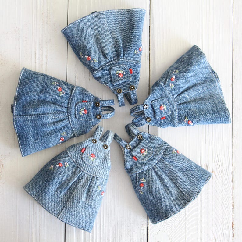 May include: Five denim dresses with embroidered floral details are arranged in a circular pattern. Each dress features a bib pocket and adjustable straps. The dresses are a light blue color and are set against a white wooden background.