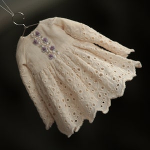 May include: A delicate, off-white dress with a scalloped hem and embroidered floral details. The dress has a long sleeve and a gathered skirt.