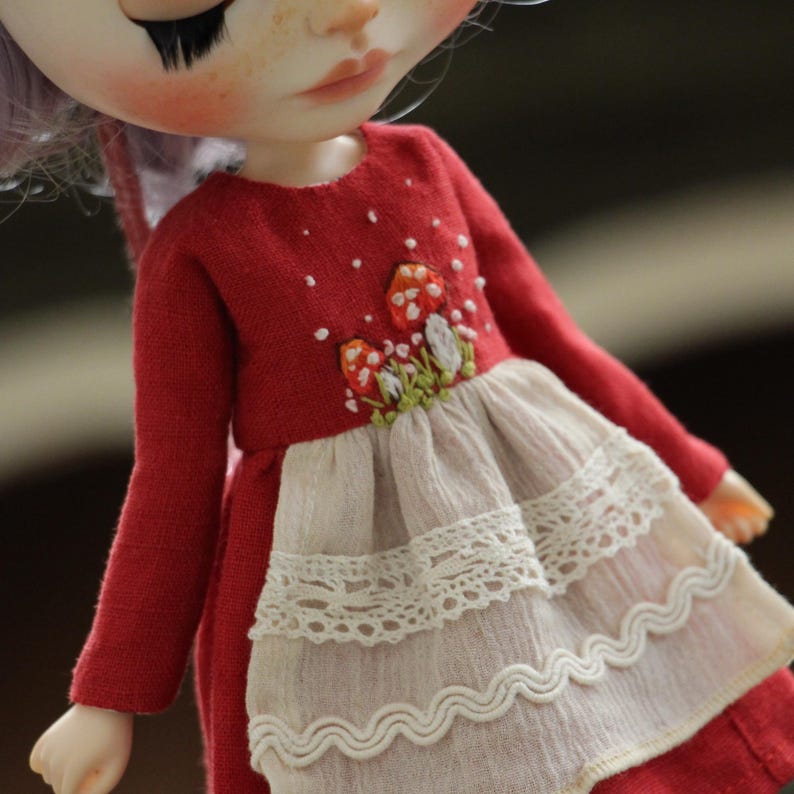 May include: A red doll dress with long sleeves and a white lace trim. The dress has embroidered mushrooms in shades of red, orange, and green.