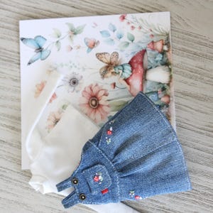 May include: A doll-sized denim overall dress with floral embroidery and a white long-sleeved shirt are displayed on a floral-themed card. The card features illustrations of butterflies, flowers, and a fairy. The overall dress has small pockets and adjustable straps.
