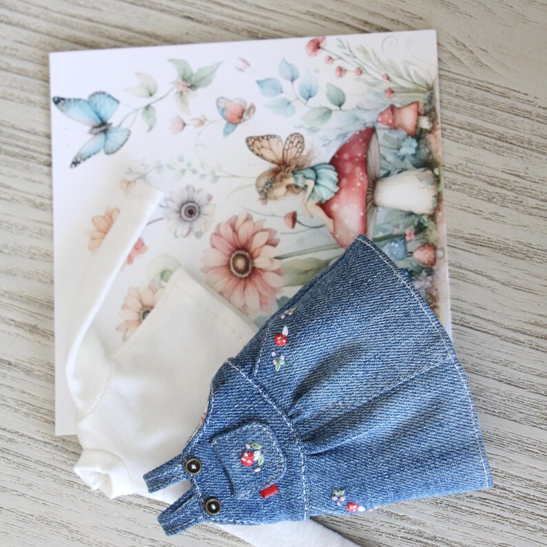 May include: A doll-sized denim overall dress with floral embroidery and a white long-sleeved shirt are displayed on a floral-themed card. The card features illustrations of butterflies, flowers, and a fairy. The overall dress has small pockets and adjustable straps.
