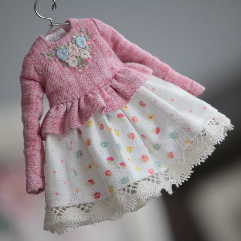 May include: A pink knitted sweater with a floral embroidery design, worn over a white dress with a floral print and white lace trim.