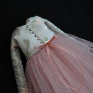 May include: A doll's dress featuring a light blue floral patterned bodice with silver bead buttons and long sleeves. The dress has a pink tulle skirt and a matching pink ribbon bow at the waist. The background is black.