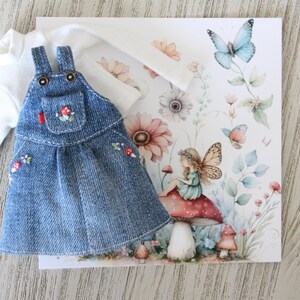 May include: A denim overall dress with embroidered mushroom details, paired with a white long-sleeved shirt. A floral and butterfly illustration is in the background, featuring a fairy sitting on a mushroom.